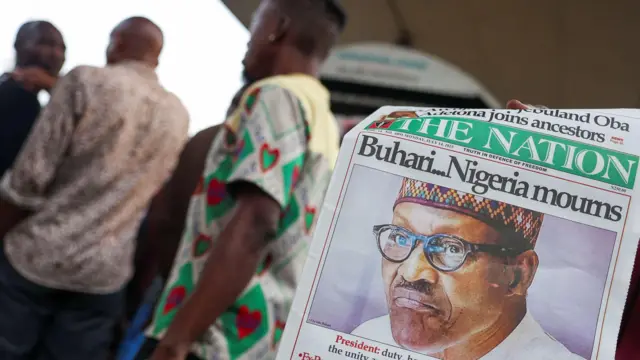 Une personne montre un journal avec un titre annonçant la mort de Muhammadu Buhari. Trois hommes apparaissent à l'arrière-plan de l'image, flous.