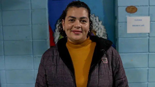 Ana Gabriela Lima, souriante à l'objectif et vêtue d'une veste marron, se tient devant un mur bleu.