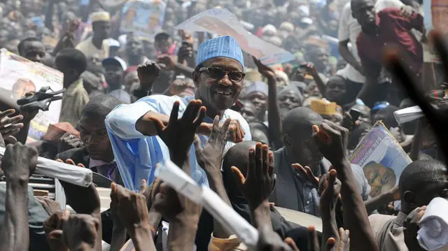 Lors d'un rassemblement plein à craquer, les partisans se pressent pour toucher Buhari, qui se tient au-dessus de la foule en souriant. Il porte un grand boubou et un chapeau bleus.