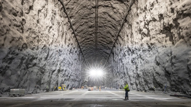 Arka planda turuncu tulumları olan işçiler, büyük beyaz bir mağarada küçük böcekler gibi gözüküyor. Arkada iki parlak ışık kaynağı var. Öndeyse parlak yeşil bir ceket giyen bir kişi, elinde bir laptopla çalışıyor