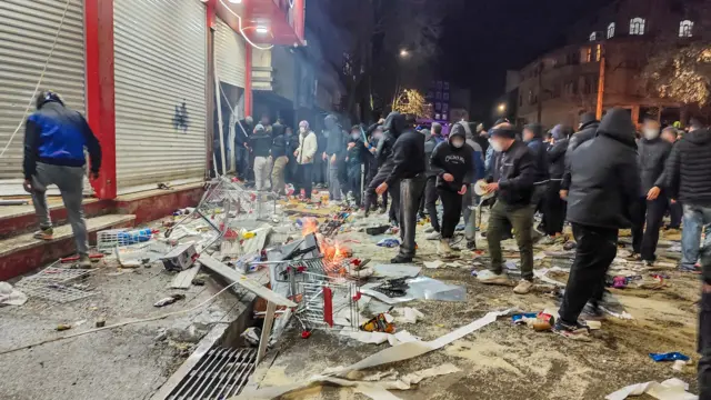 A group of pipo wey wear face masks and hoods gada around burning remains outside closed shops for Kermanshah on Thursday.