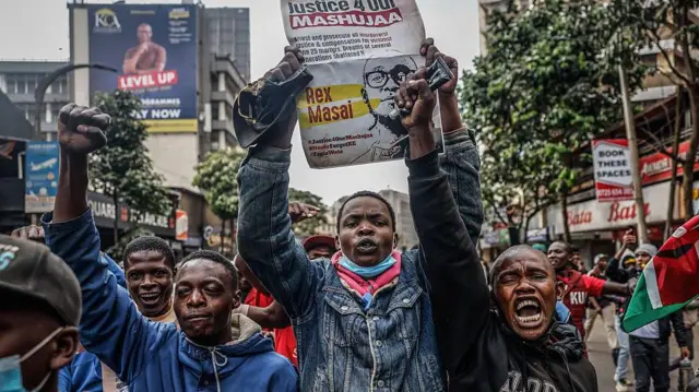 Demonstrators march for downtown Nairobi on June 25, 2025 during a day of protest to mark di first anniversary of di parliament protest. A young man holding a placard, anoda man raise im hand wit mouth open, oda men for di background in protest