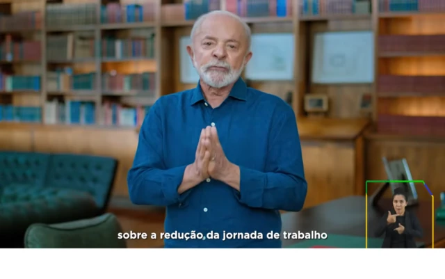 O presidente Luiz Inácio Lula da Silva durante pronunciamento televisivo pelo Dia do Trabalhador, em 1º de maio de 2025. Ele veste camisa azul com as mangas compridas dobradas e tem as mãos unidas junto ao peito. Uma legenda em letras brancas diz "sobre a redução da jornada de trabalho" e há uma tradutora de Libras no canto direito da imagem
