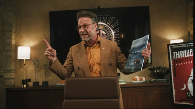 Un hombre habla con los brazos abiertos y un dossier en la mano derecha en lo que parece ser un despacho. A su izquierda se ve un cartel con el texto "Thriller movies".