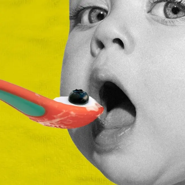 A baby in black and white opens its mouth and looks into the camera while it is fed a spoonful of yoghurt and with a blueberry on top. The background is yellow and the spoon is orange and green.