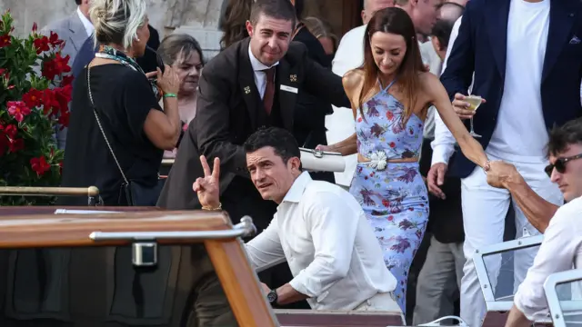 Орландо Блум в белой рубашке изображает рукой знак мира и садится в лодку.