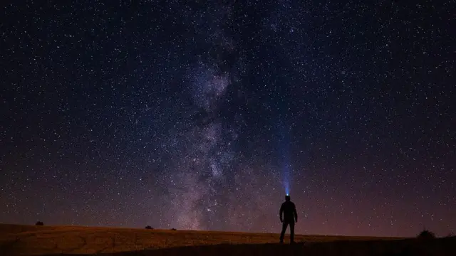 Başında ışık olan bir kişinin küçük silüeti, sayısız yıldızla dolu uçsuz bucaksız gece gökyüzüne bakıyor; ortada ise Samanyolu galaksisi yukarı doğru uzanıyor.
