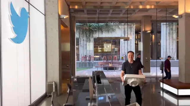 Elon Musk menggotong wastafel dapur ke dalam kantor X, dulu bernama Twitter. Di dinding terdapat logo Twitter yang lama.
