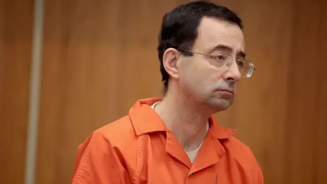 Larry Nassar, vu de la taille jusqu'au cou, portant des lunettes à monture carrée avec des branches métalliques et une combinaison orange, regarde vers sa gauche.