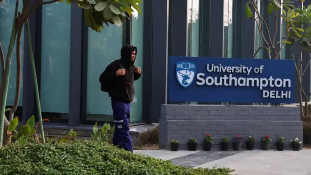 A man wearing a brown hoodie and blue trousers passes by the entrance to the campus. On his side is a board which reads University of Southampton Delhi. There are trees and shrubs on the side and a glass building in the backdrop.