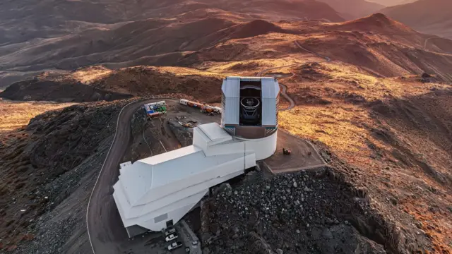 An aerial view of a huge telescope seen through its open housing, at the top of a large, white building perched on the edge of a rocky mountaintop. There is a road around the observatory and some cars at the bottom, with the mountainous landscape glowing golden brown in the sunset.