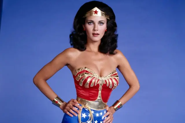 Lynda Carter con su característico disfraz de Wonder Woman. El atuendo tiene un top rojo y dorado, con pantalones cortos azules con estrellas blancas. Tiene una corona triangular dorada en la frente con una estrella roja, puños plateados con estrellas rojas y una cuerda dorada llamada lazo de la verdad que sale de su cinturón.