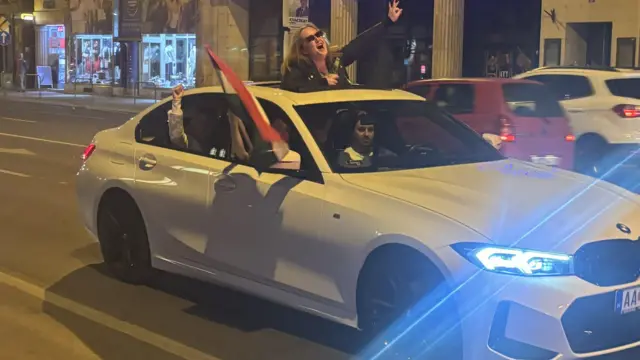 흰 차 밖으로 헝가리 국기를 흔들고 있고, 한 여성은 선루프를 통해 밖으로 나와 기쁨을 만끽하고 있다