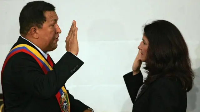 Ông Hugo Chavez giơ tay phải về phía bà Flores, người cũng làm điều tương tự trong một nghi thức dường như là lễ tuyên thệ nhậm chức. Cả hai đều mặc vest tối màu và có vẻ mặt nghiêm nghị.