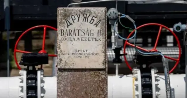 Фрагмент нафтопровідної інфраструктури з вентилями та трубами. У центрі — кам’яна табличка з написами «ДРУЖБА» та угорською «BARÁTSÁG kőolajvezeték» («нафтопровід “Дружба”»), із датою будівництва 1970–72. Це позначення радянського трубопроводу «Дружба».