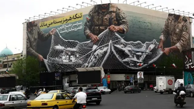 交差点に面した建物の壁面に大きく掲げられたポスターに、イランの軍人3人が網で、米軍の戦闘機や艦船や魚雷などをからめとっている様子が描かれている。交差点では乗用車やトラックやバイクに乗った人達が行きかっている。