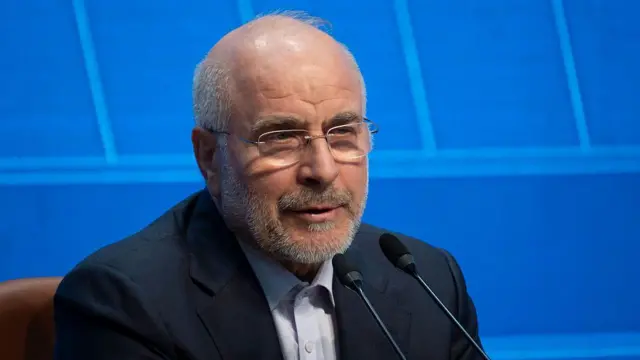 Mohammad Bagher Ghalibaf wearing a white shirt and dark blue blazer, speaking into a microphone against a blue background.