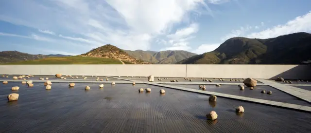 Patio con lámina de agua y rocas dispersas, enmarcado por un muro recto y un paisaje de montañas bajo un cielo despejado.