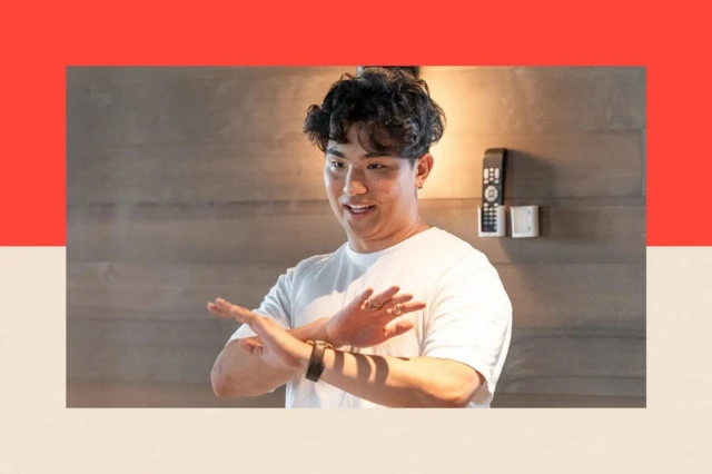 Мужчина в белой футболке с азиатской внешностью. У него кудрявые темные волосы, на левой руке у него часы, на правой кольца на пальцах