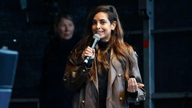 Kahverengi deri ceketli ve uzun kumral saçlı bir kadın elinde bir mikrofon tutuyor