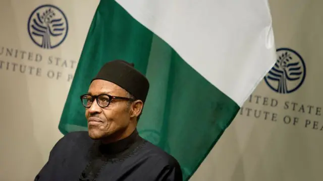 Muhammadu Buhari est assis devant un drapeau nigérian et des panneaux indiquant « United States Institute of Peace ». Il porte un boubou noir, un chapeau noir et des lunettes.