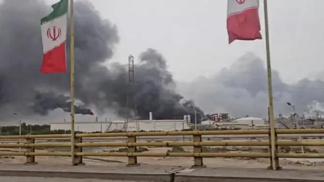 Дым поднимается в воздух после ударов по нефтехимической зоне Махшахр в округе Бандар-Махшахр провинции Хузестан, Иран. На переднем плане у дороги на флагштоках развеваются два иранских флага, а позади них виден подвергшийся удару завод