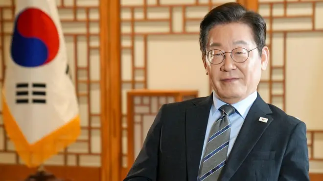 李在明總統身穿黑色西裝,坐在首爾辦公室的椅子上。他身後是兩面國旗。