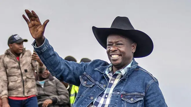 Rigathi Gachagua yambaye ijaketi y'ikoboyi y'ubururu, n'ingofero y'urugara y'umukara imenyerewe ku bashumba b'inka, arimo gupepera abamushyigikiye, mu mwaka wa 2025.