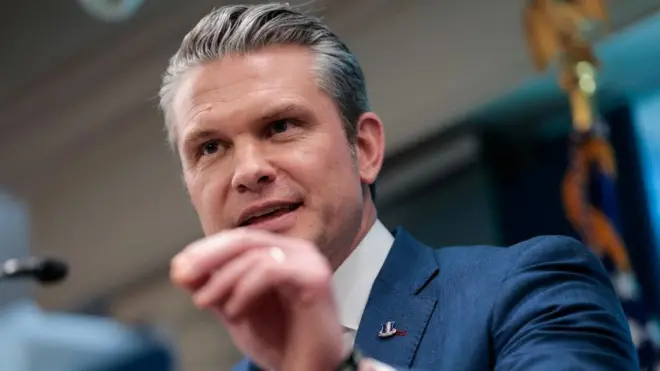 Head and shoulders pic of Pete Hegseth at the podium of the Pentagon briefing room. He's wearing a well-tailored petrol blue suit with a blue and pink tie on a white shirt. His left hand is raised and his thumb and index finger joined as if emphasising a point