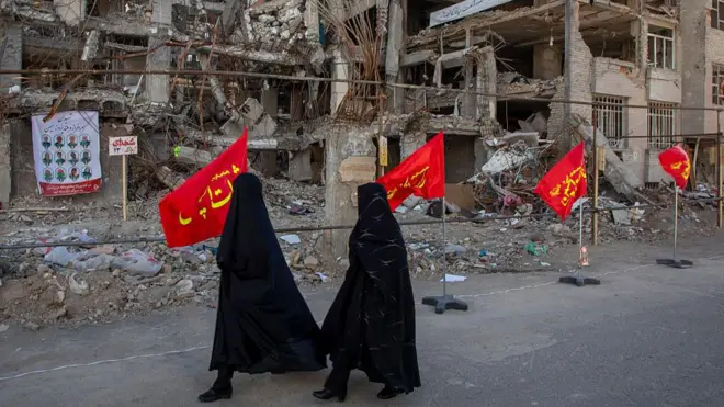 Two veiled women in long black robes walk past red flags planted in front of a heavily damaged concrete buildings destroyed in a joint attack by Israel and the United States on 6 April 2026, in Tehran, Iran.
