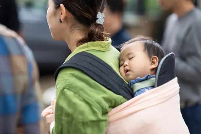A woman in Japan walks down a street carrying a baby in a sling on her back. She is wearing her hair in a bun and a green jacket, and the baby is asleep. 