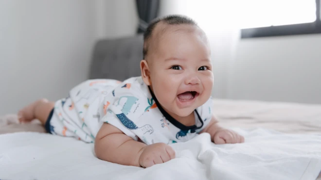 A smiling baby boy lying on his tummy on a bed ඇඳක් මත වැතිර සිටින පිරිමි ළමයෙක්