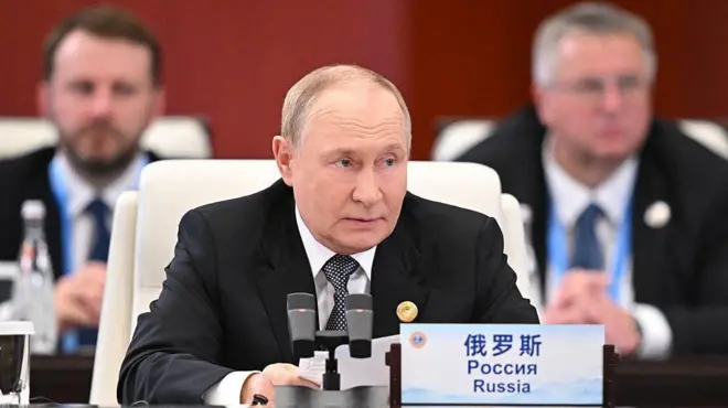 Vladimir Putin, wambaye ikositimu yijimye na karuvati y'umweru n'umukara, yicaye imbere y'ameza mu nama, arimo kuvugira mu ndangururamajwi. Icyapa kiri imbere ye ku meza cyanditseho "Uburusiya" mu Gishinwa, mu Kirusiya no mu Cyongereza.