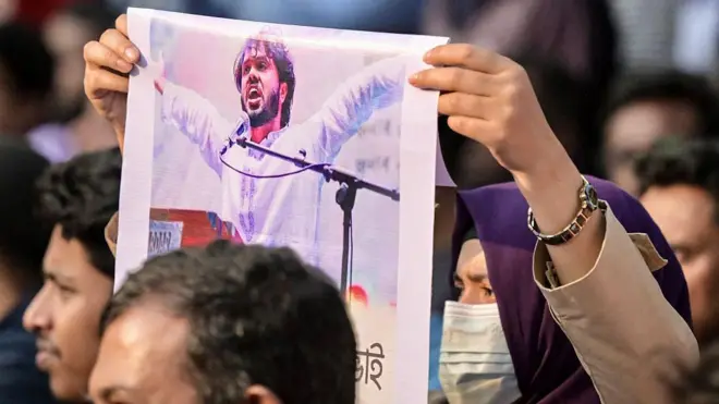 မျက်နှာဖုံးတပ်ထားတဲ့ အမျိုးသမီးတစ်ဦးက ကျောင်းသားဆန္ဒပြအဖွဲ့ Inqilab Mancha ရဲ့ ထိပ်ပိုင်းခေါင်းဆောင် ရှာရစ်ဖ်အော့စ်မန်ဟာဒီ ရဲ့ ဓာတ်ပုံကို ကိုင်ဆောင်ထားပါတယ်။