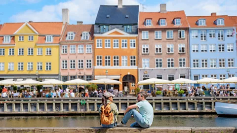 Two people sit by the canal in Copenhagen in front of colourful buildings (Credit: Getty Images)