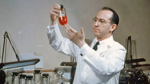 Dr Jonas Salk stands in a lab holding up a beaker with a bright orange liquid filling a quarter of it, and lots more beakers full of the liquid underneath (Credit: Alamy)