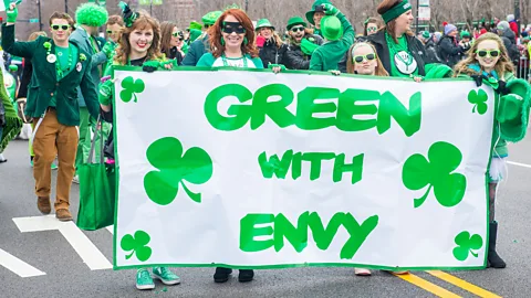 At a parade, four young women dressed in green hold a banner that reads "Green with Envy", with six clovers spaced around the words (Credit: Alamy)