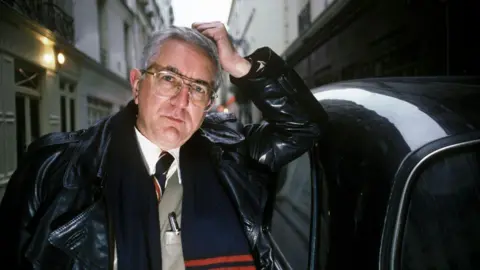 Len Deighton is seen staring into camera. He is leaning on the roof of a black London tax with his elbow and has his hand on his head.
