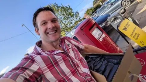 A man with brown hair in a red and white checked shirt takes a selfie standing next to a post box. He has a box under his arm containing stacks of black envelopes. It is a sunny day and he is smiling.