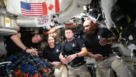 NASA Inside a cramped spacecraft cabin, five astronauts float side by side in weightlessness. They wear matching black T‑shirts with a small mission patch on the chest and pale trousers with Velcro straps. Their faces are blurred, but their relaxed body language suggests they are chatting to camera. Behind them, every surface is crowded with white padded panels, cables, pipes and equipment. Above their heads, American and Canadian flags are pinned to a storage bag, with a colourful “America 250” sign between them. To the left, a netted bundle of blue and orange bags is tethered down, preventing it from drifting. 