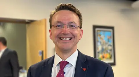 BBC Deputy Kirsten Morel, a man with short brown hair and glasses with no frame. He is staring directly at the camera and smiling. He is wearing a white shirt, maroon tie and a navy blue blazer. He has a red lapel badge. Behind him, out of focus, a colourful piece of artwork is visible on the wall, in a dark frame.