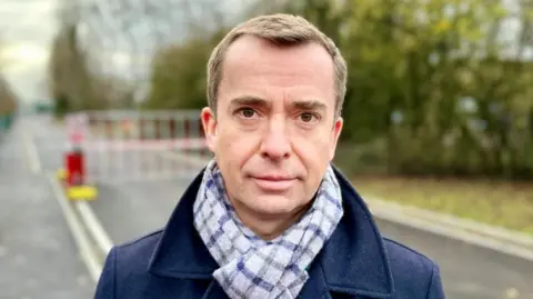 A man is looking straight at the camera. He is wearing a checked scarf and a blue coat.