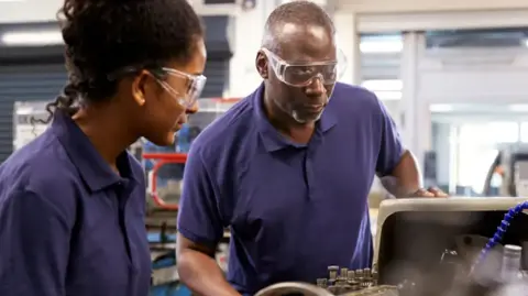 A man and a woman stand in a workshop looking at a equipment while wearing goggles. They both have purple T-shirts on and equipment can be seen behind them.