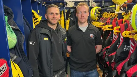 Two men wearing black tops stand inside a lifeboat station. Equipment, uniforms and helmets are seen in the background