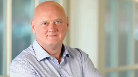 FICO Campbell Scott staring straight at the camera. He is bald. He has blue eyes. He is wearing a light blue shirt with dark-coloured checks on it. He has his arms folded and is standing in front of a blurred background.