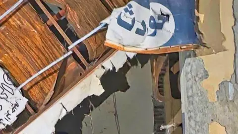 Ruins of walls and a ceiling at el-Daein Teaching Hospital. A blue and white sign with the word reading 'Accidents' in Arabic is seen hanging in tatters.