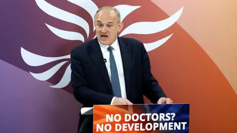 Liberal Democrat leader Sir Ed Davey gives a speech from behind a podium reading: "No doctors? No development". He is standing in front of a purple and orange backdrop with a white Liberal Democrat logo of a bird.