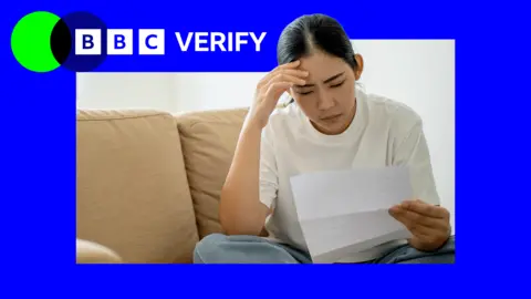 A woman with dark hair in a ponytail, sitting on a beige sofa and looking at a bill, with her hand on her head 