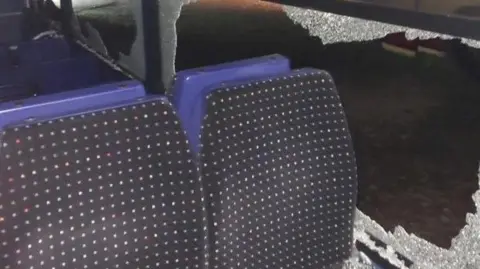 The interior of a coach which has had all of its windows smashed. There are shards of glass all over the dark patterned fabric seats. It is dark outside.