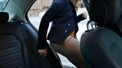A woman stepping in to the back seat of a car from a city street, wearing a navy blazer and beige trousers